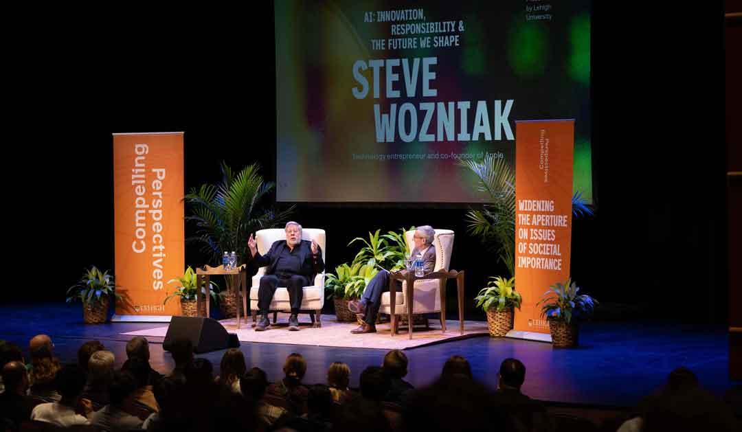 Steve Wozniak speaking on stage with audience, surrounded by plants and banners.