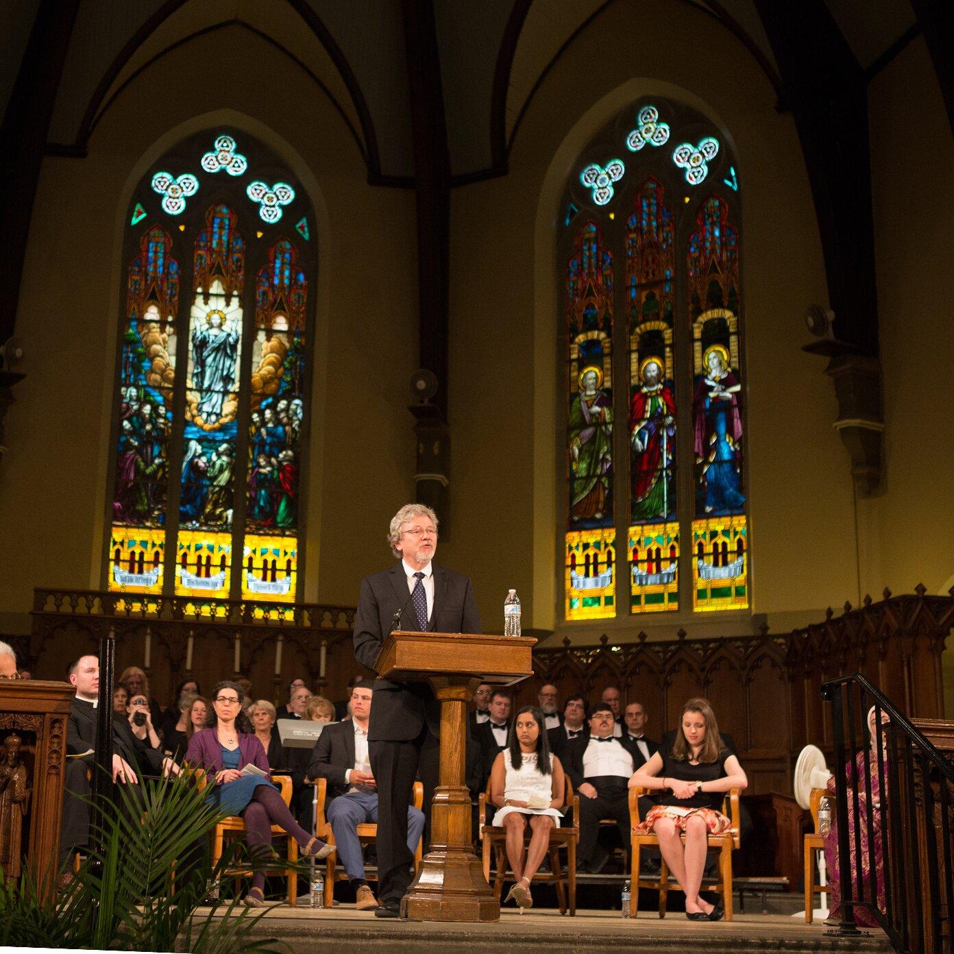 Man speaking from a podium in a hall with large stained glass windows and an audience.