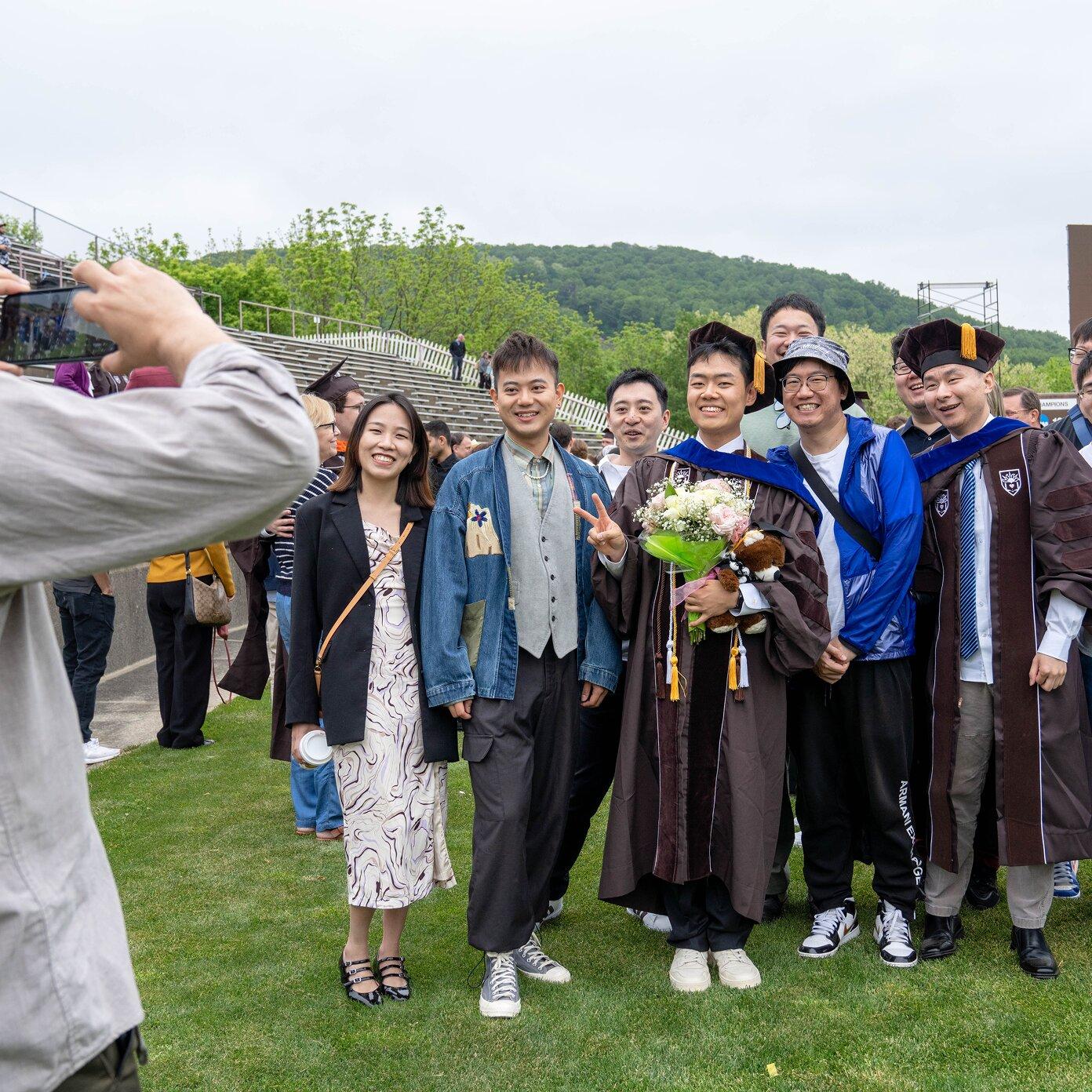 Graduate in academic gown holding flowers, smiling with family and friends for a photo.