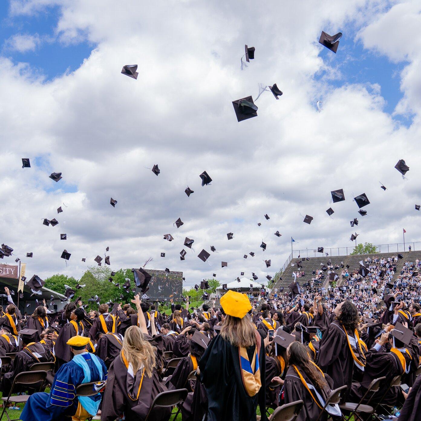 Graduates in brown gowns toss caps into the air, celebrating under a cloudy sky at a stadium.