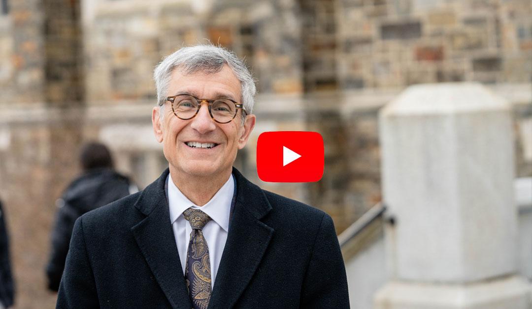 Smiling man in a black coat stands outside a stone building, with a YouTube logo in the corner.