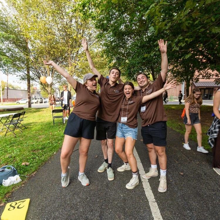 Four young adults in brown shirts cheer with arms raised.