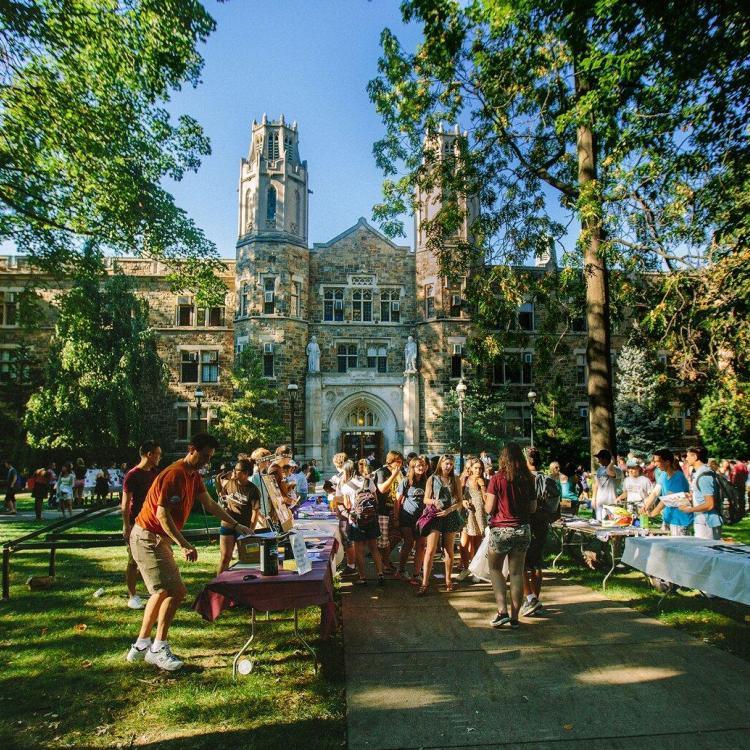 Students gather outside a large stone building with towers on a sunny day.