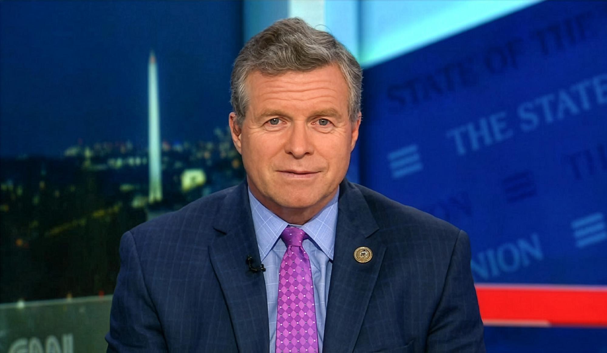 Man in blue suit and purple tie in a studio with Washington Monument background.