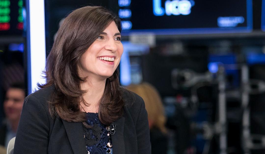 Smiling woman with dark hair in a black blazer against a blurred financial background.
