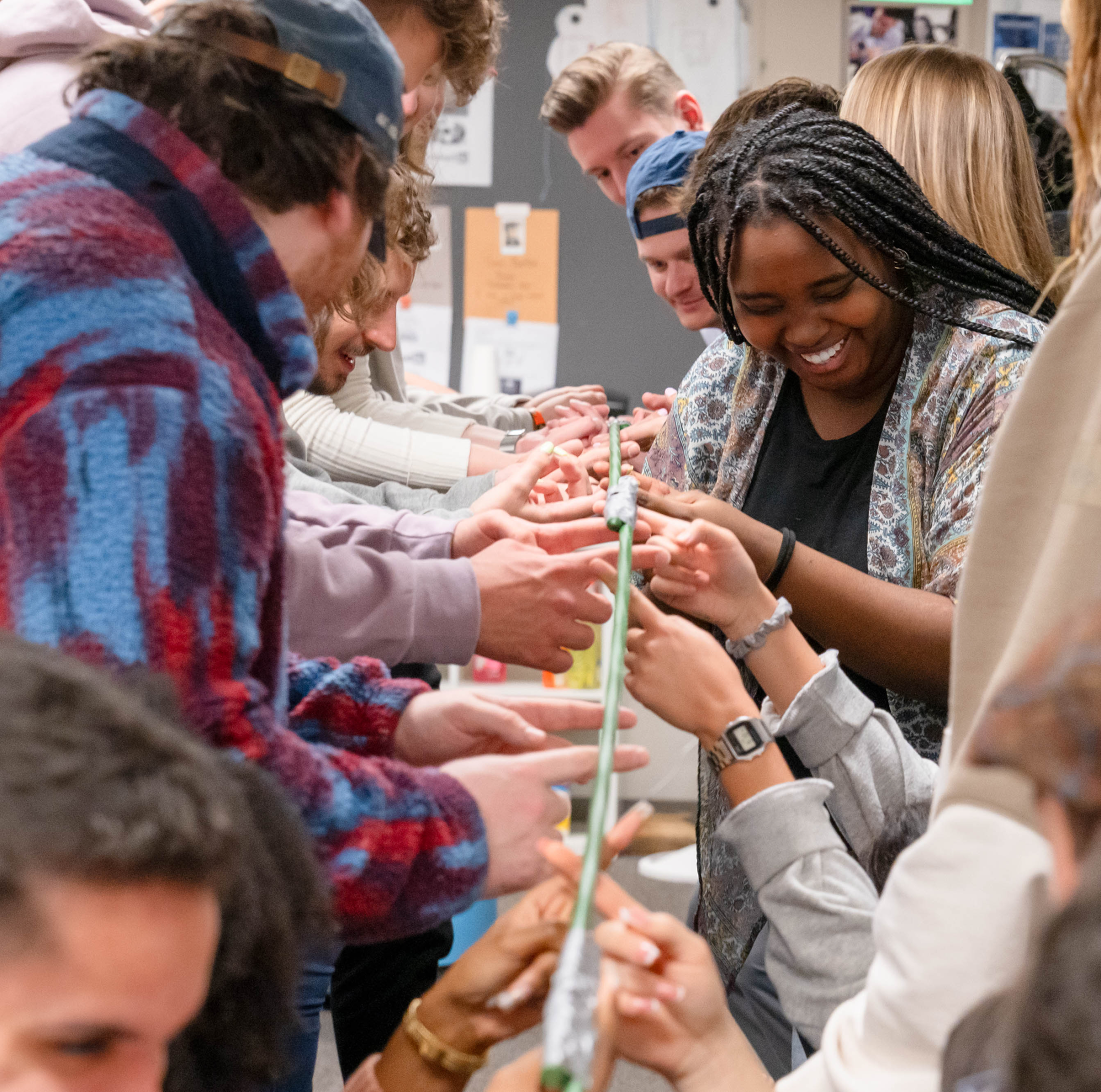 A diverse group of people stand in two lines facing each other, collaborating and smiling as they balance a long, thin object together, likely participating in a team-building activity indoors.