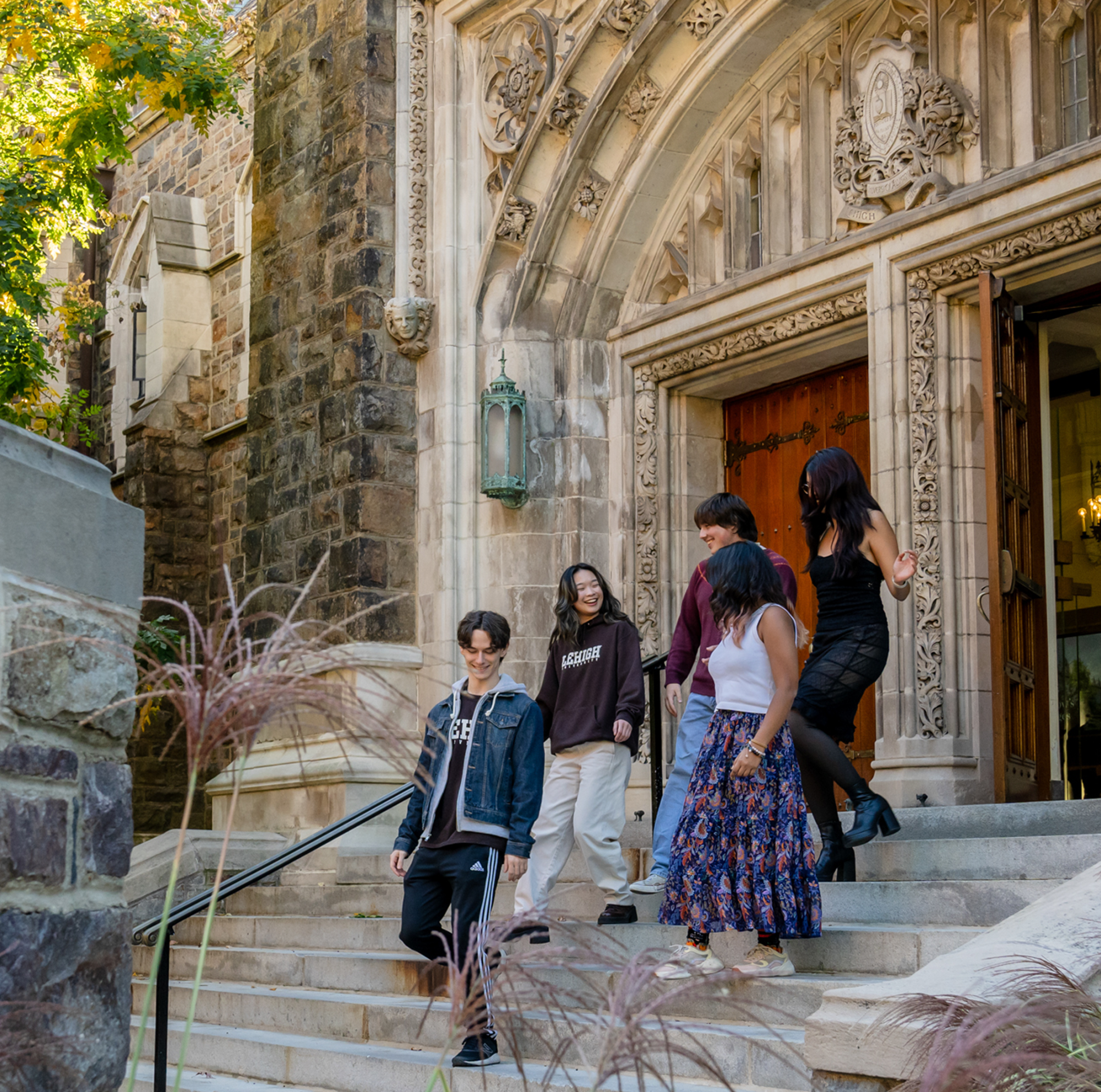 Four students walk down the stone steps of a historic building with ornate carvings around the arched entrance, surrounded by greenery on a sunny day.