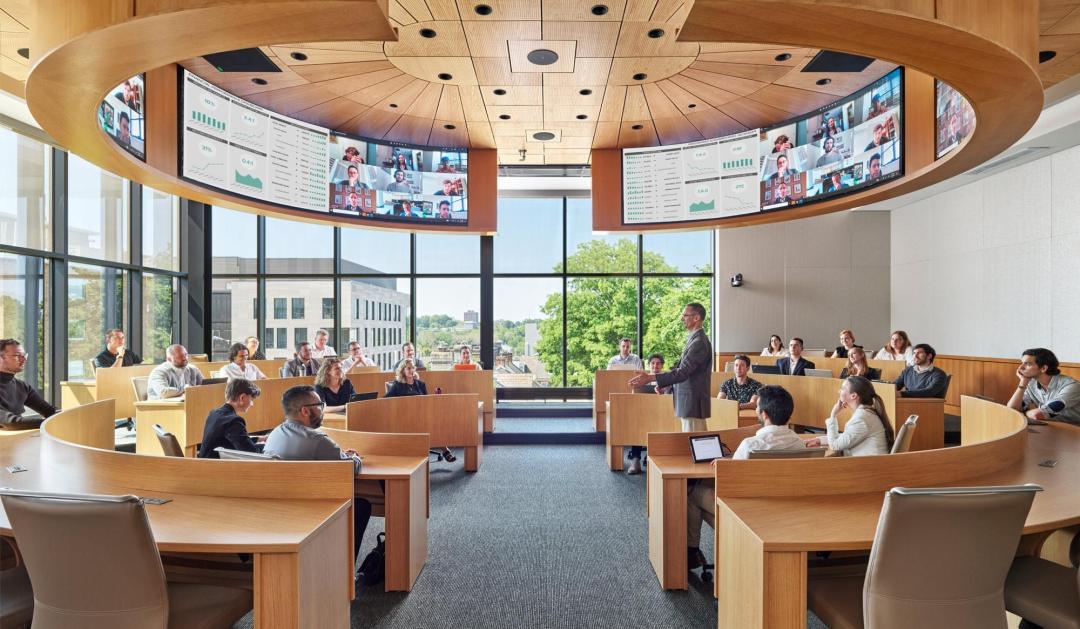 Professor lectures students in a modern, semi-circular classroom with large curved display screens.