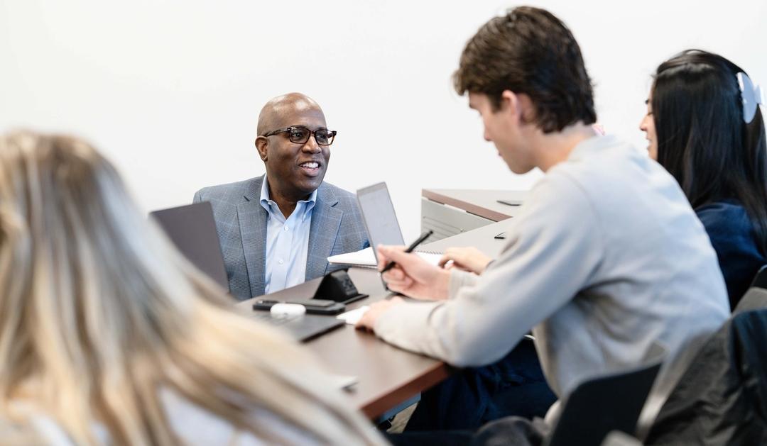 A professor sitting with students, all engaged in a discussion around a conference table.