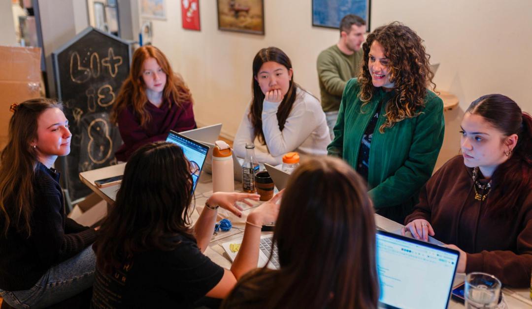 Group of students collaborating around a table with laptops as Kate Jackson sits in the middle and speaks.