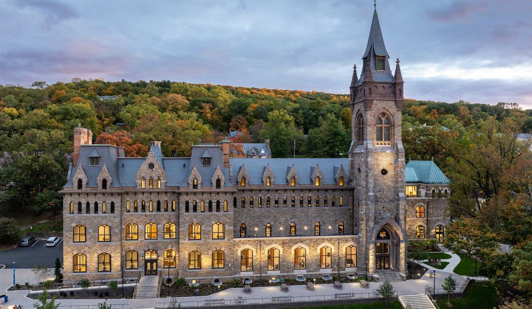 Photograph of a lit Gothic stone building and tower at dusk against a wooded hillside.