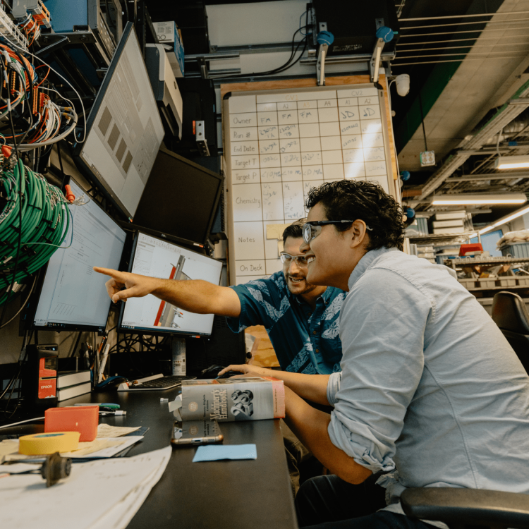 Two people sit at a desk surrounded by computer monitors and technical equipment in a modern lab or office, collaborating as one points at a screen while the other types on a keyboard.