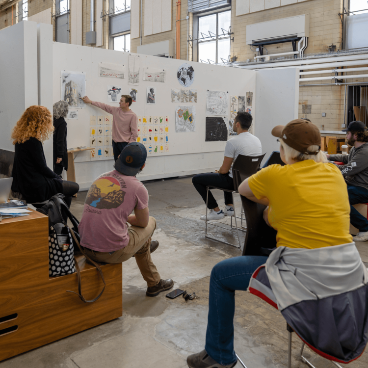 A teacher stands at a whiteboard, explaining a concept to a group of seated students in an art classroom. Various sketches are displayed on the walls, and the students focus attentively on the lesson.