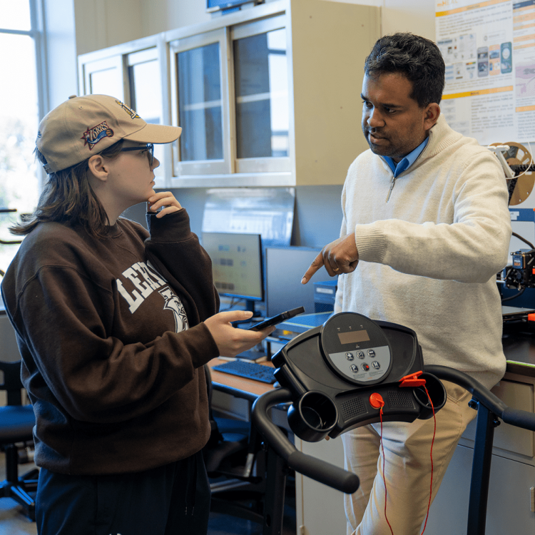 A man in a white sweater explains a piece of lab equipment to a woman in a brown sweatshirt and cap inside a laboratory, with computers and shelves visible in the background.