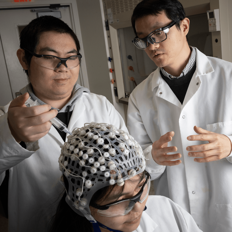 Three scientists in lab coats and safety glasses observe a colleague wearing an EEG cap, discussing and examining data in a laboratory setting.