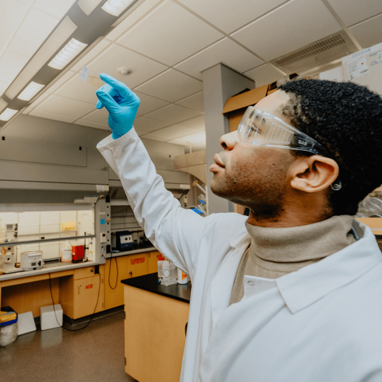 A scientist wearing safety goggles and blue gloves points upward in a laboratory, surrounded by lab equipment and shelves.