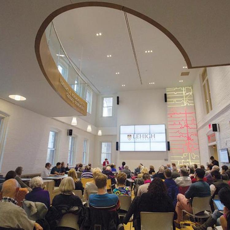 A large group of people sit facing a presenter and a projection screen in a modern, well-lit conference room with high ceilings and round windows.