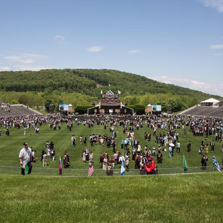 Outdoor graduation with a crowd on a field, stage in background, and green mountains.