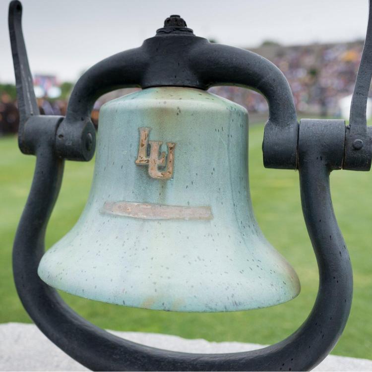 Bronze bell with a green patina and an 'LU' logo, mounted on a black metal stand.