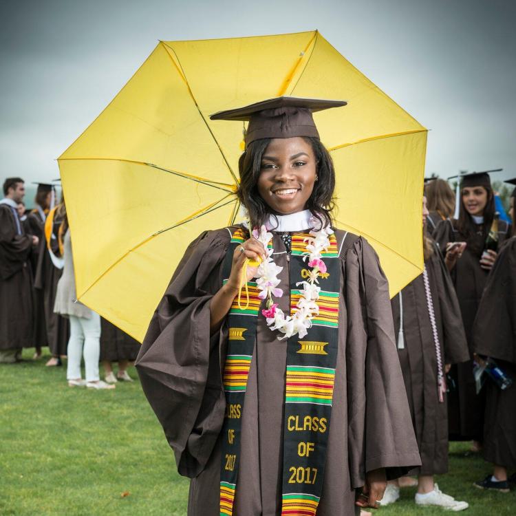 Smiling graduate in brown gown and lei holds a yellow umbrella.