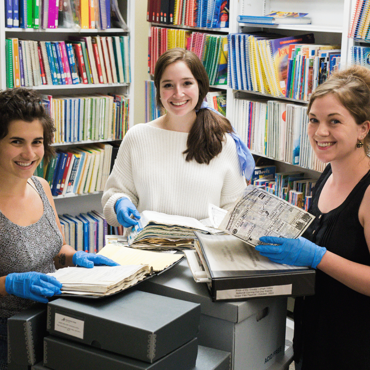 Three smiling women wearing blue gloves examine archival documents in a library.