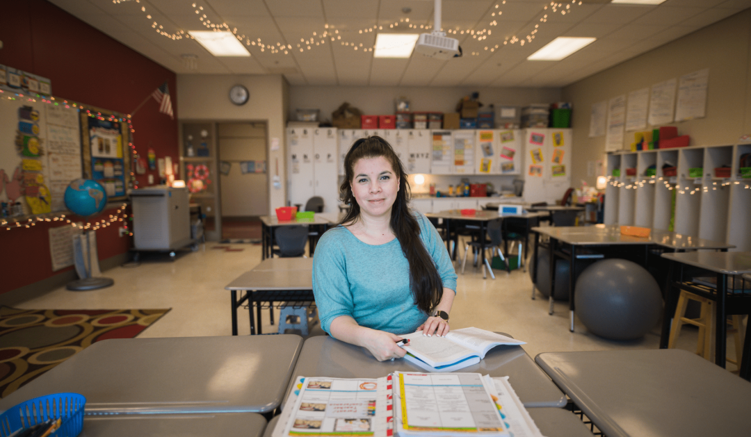 A woman with long dark hair sits at a desk with open books in a colorful, well-lit classroom decorated with educational materials and string lights.