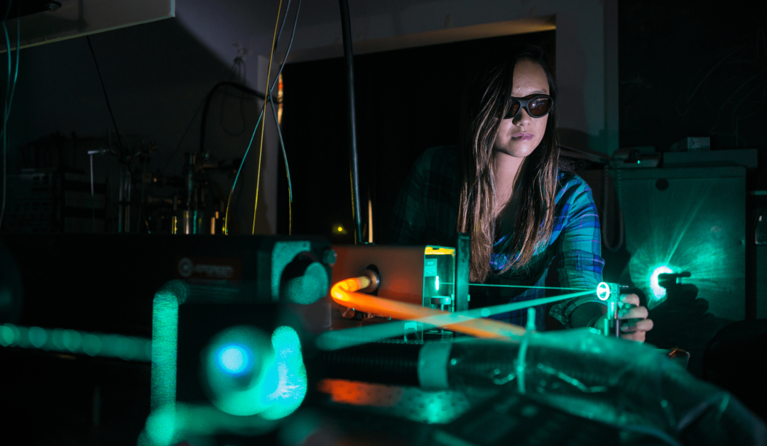 A person wearing safety glasses operates scientific equipment in a dark laboratory, with glowing green and orange laser beams illuminating the scene.