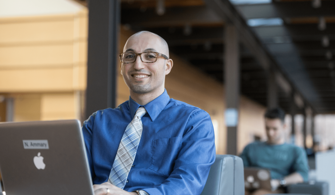 A man wearing glasses, a blue shirt, and a striped tie sits at a table working on a laptop in a bright, open indoor space. Another person is seated in the background, also using a laptop.