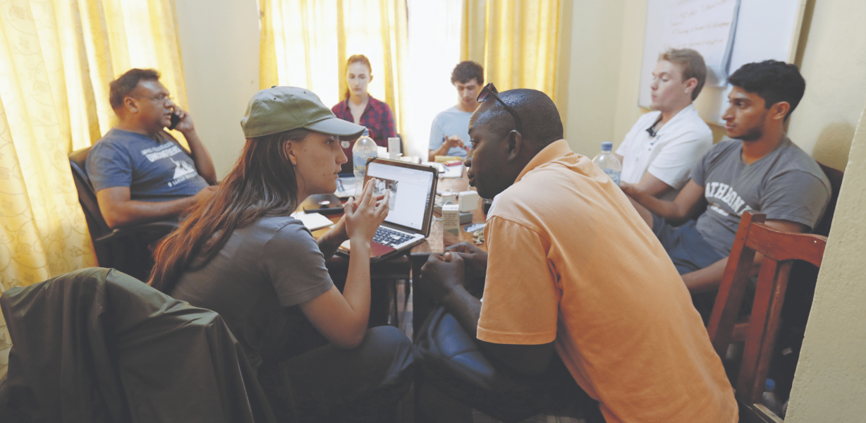 A group of eight people sit around a table in a small, sunlit room, engaged in discussion. Some are talking, while others listen attentively; a laptop and notepads are visible on the table.