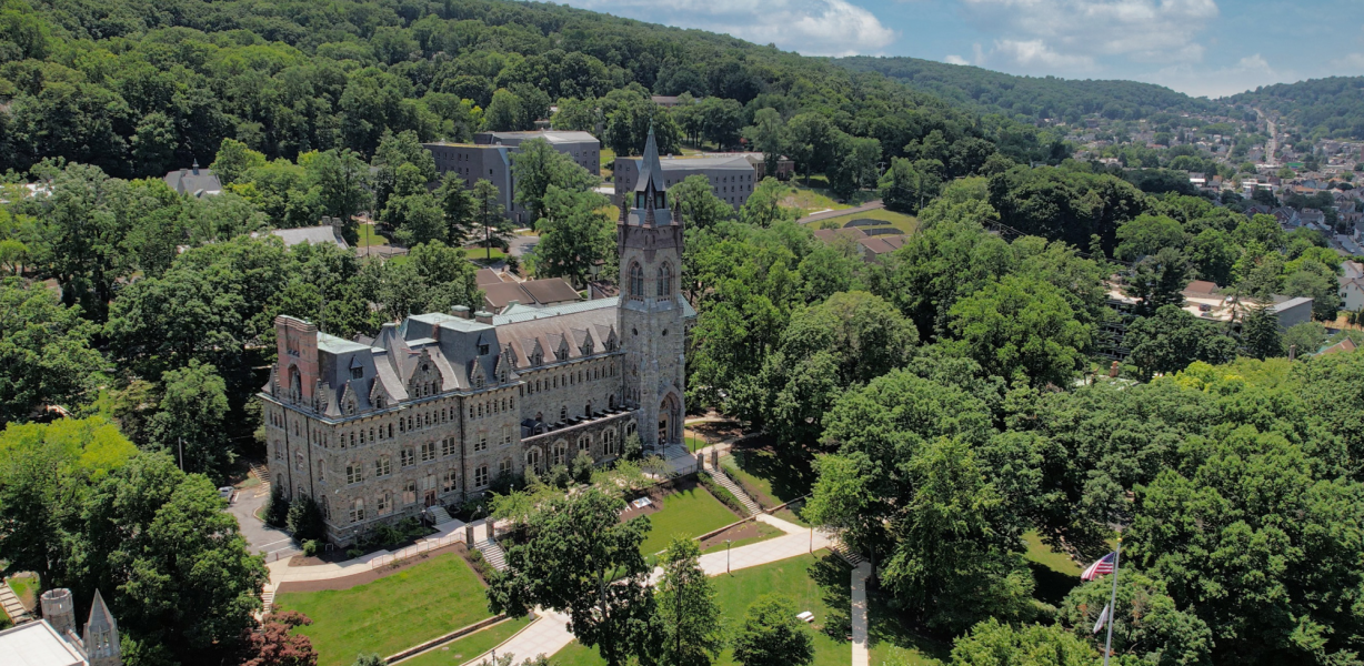 Aerial view of a large, historic stone building surrounded by trees and greenery, with a hillside and blue sky in the background on a sunny day. Pathways and lawns are visible around the building.