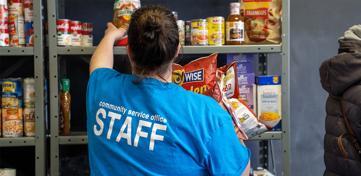 Volunteer in blue shirt with "STAFF" written on back reaches for a jar on a shelf of canned goods.