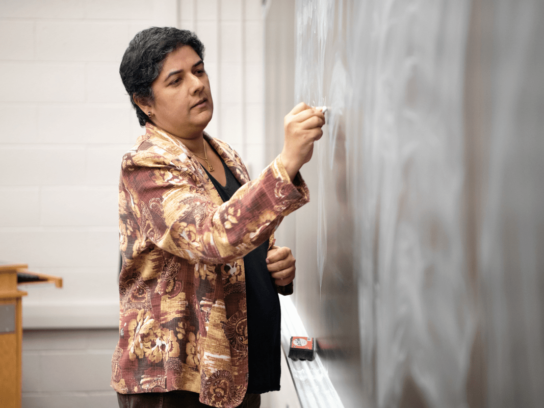 A person wearing a patterned jacket stands at a chalkboard, writing. A wooden podium is visible in the background. The setting appears to be a classroom or lecture hall.