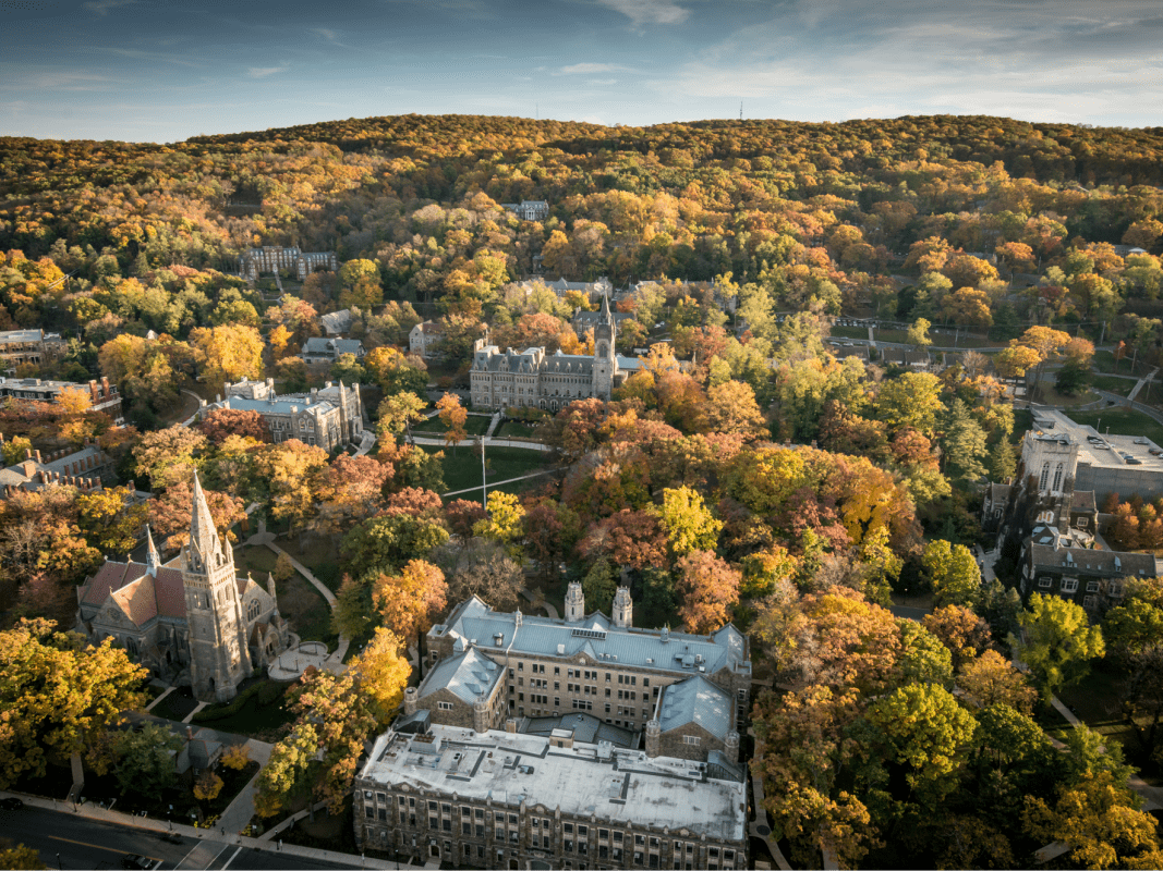 Aerial view of a college campus in autumn, with buildings surrounded by colorful trees and rolling hills in the background under a partly cloudy sky.