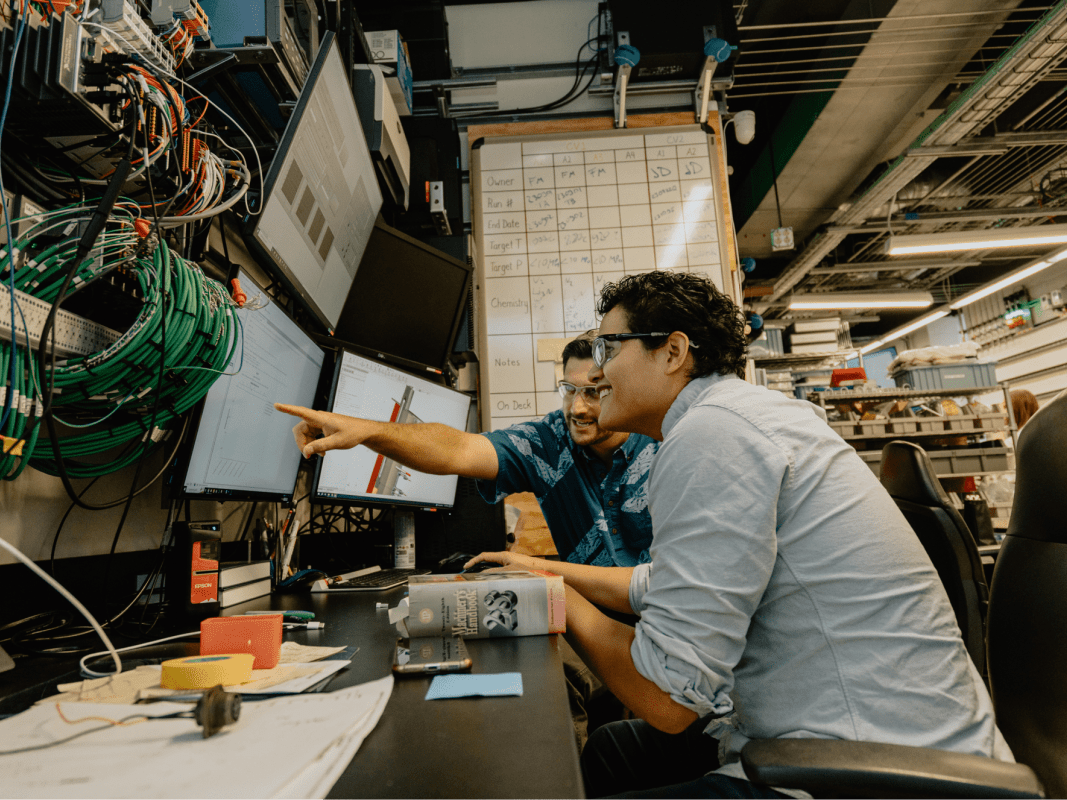 Two people sitting at a workstation with multiple monitors and electronic equipment, one person pointing at a screen while the other observes intently in a technology or engineering lab setting.