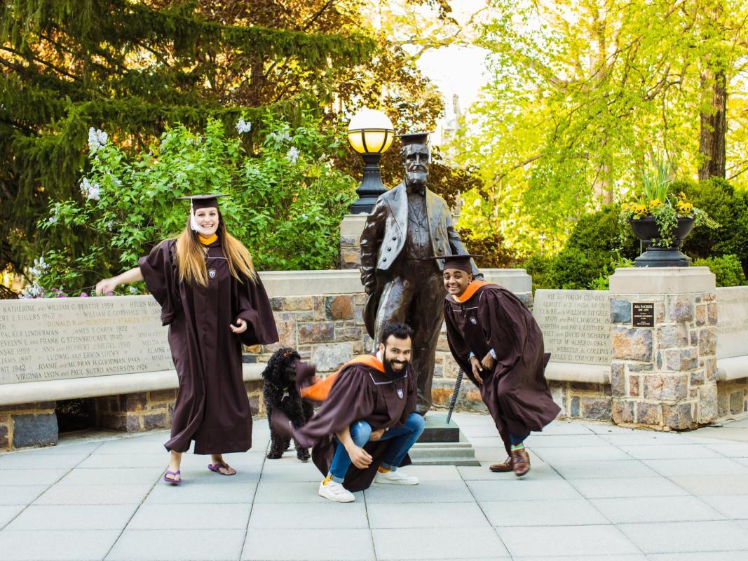 Three graduates pose playfully around a statue on a sunny campus.