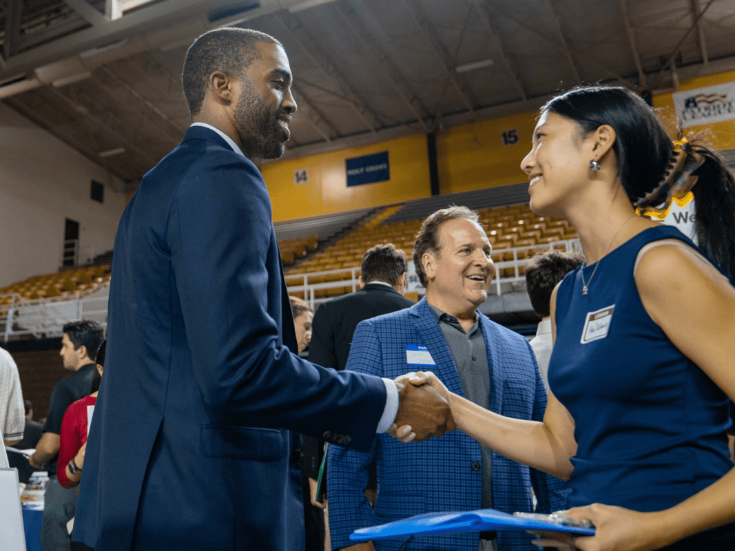 A man in a suit shakes hands with a woman in a navy blue top, both smiling.
