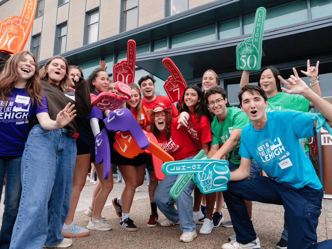 Group of excited young adults in colorful shirts and foam fingers cheer outside building.
