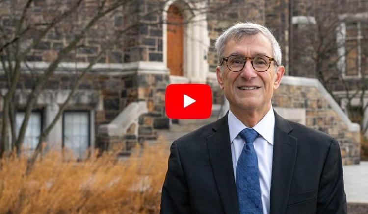 A man in a suit smiles outdoors near a stone building with winter foliage.