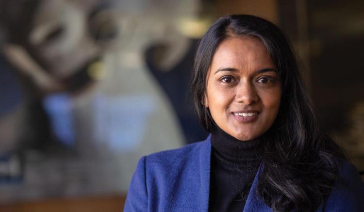 A woman with long black hair smiles, wearing a blue jacket, with a blurred background.
