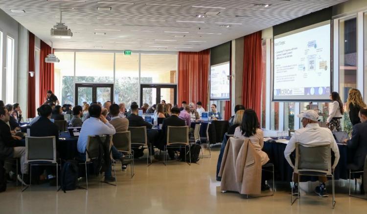 A conference room filled with attendees seated at tables, listening to a presentation.