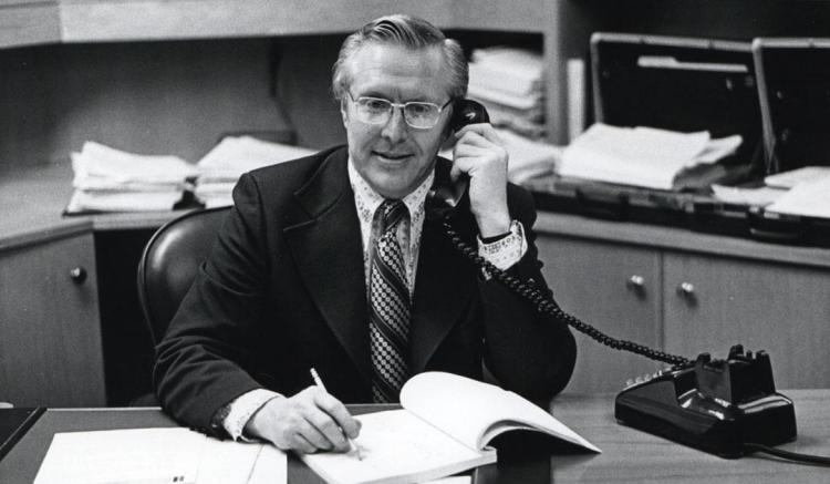 Black and white photograph: Man in suit and glasses talks on phone, writes at office desk.