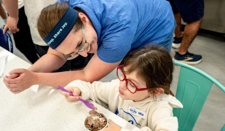 A woman leans over to watch a young girl in red glasses eat a bowl of ice cream.
