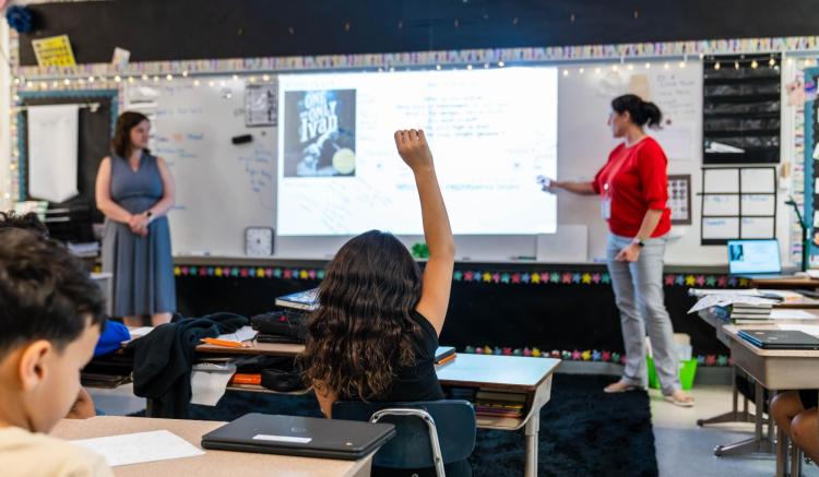 A teacher points to a projector screen as a student raises her hand in a classroom.