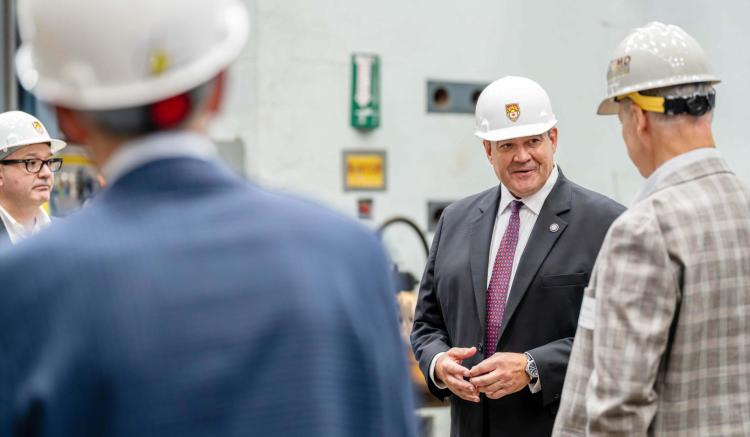 Photograph of a man in a suit and hard hat speaking to others in an industrial setting.