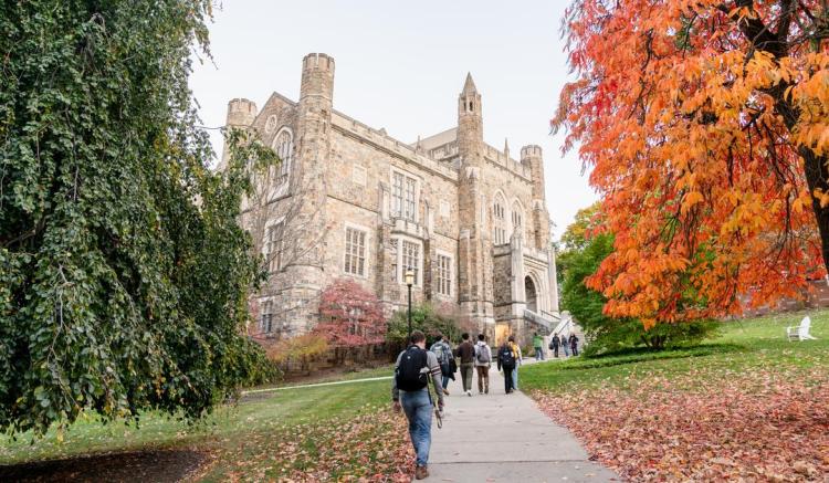Photograph of students walking toward a gothic stone building with vibrant autumn trees.